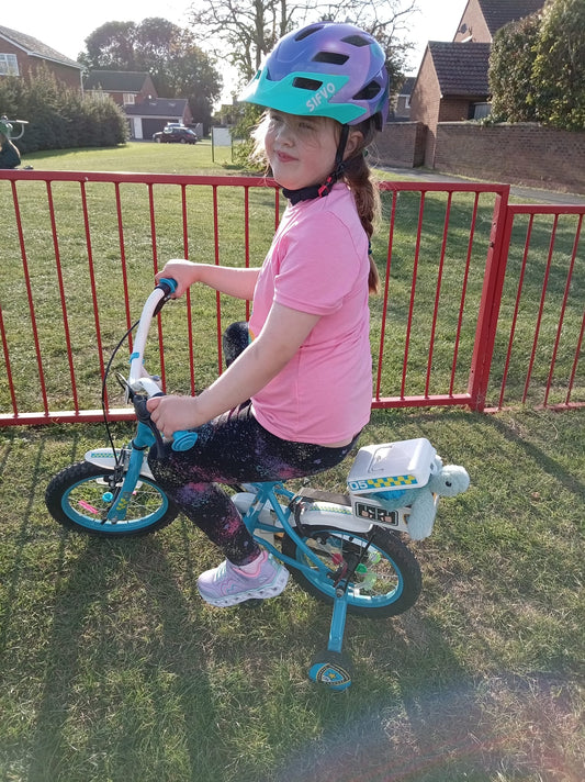 Alice on her blue police bike, wearing a pink t;shirt and purple safety helmet. Mini Sunny the seaturtle is poking out of the back of the plastic box attached to the back of the bike.
