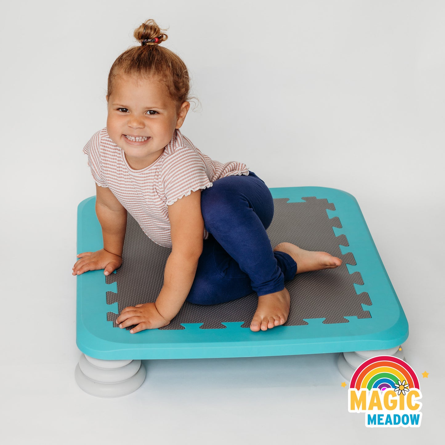 Little girl sat on blue trampoline board facing the camera smiling. Magic Meadow rainbow logo bottom right corner.