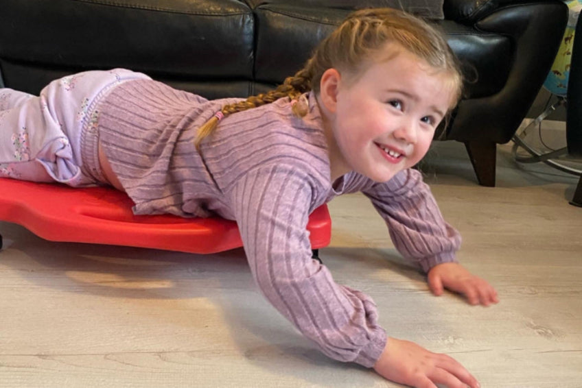 Alice laying on a red scooter board looking at the camera smiling. Her arms are outstretched with hands on the floor.
