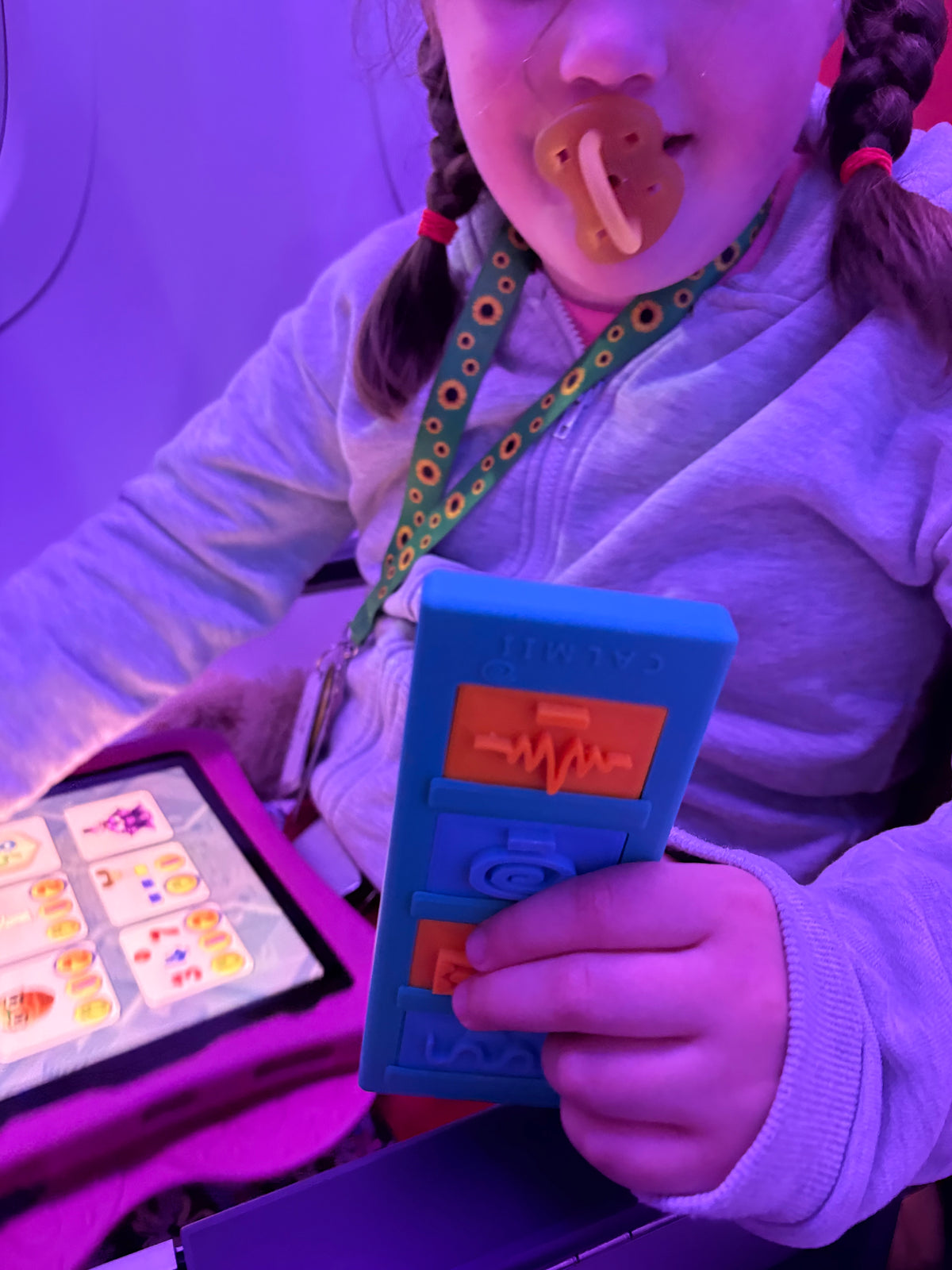 A child with braided hair and a pacifier is holding a Calmii Bar with blue and orange tiles under purple ambient lighting. She is sat in a seat on an aeroplane. A tablet is visible on her lap. She wears a sunflower lanyard.