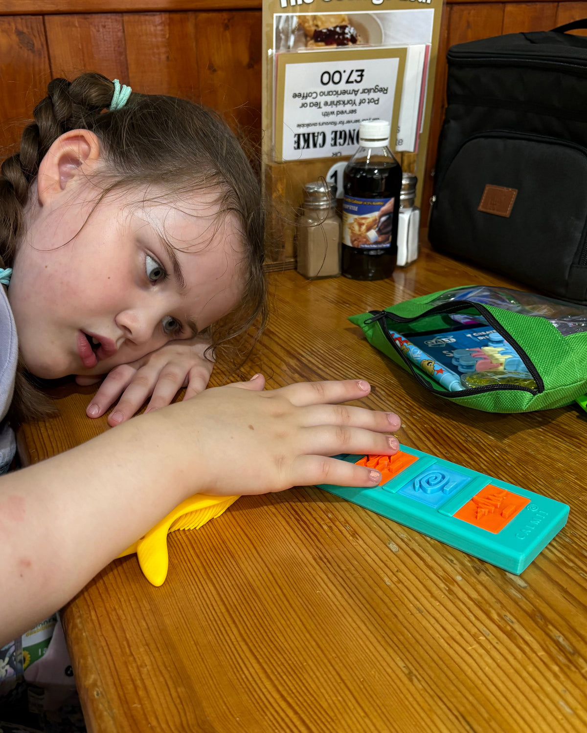 A young girl with braided hair rests her head on her hand on a wooden table at a cafe, looking at her Calmii Bar and Massage Brush. Visible items on the table include a green Grab and Go Sensory Bag, a black bag, condiment bottles, and a sign for 'The Cottage Loaf' cafe'.